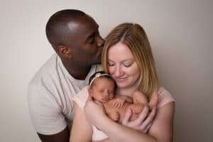 newborn photographer in rochester ny captures dad kissing mom on the head while she holds their newborn baby girl