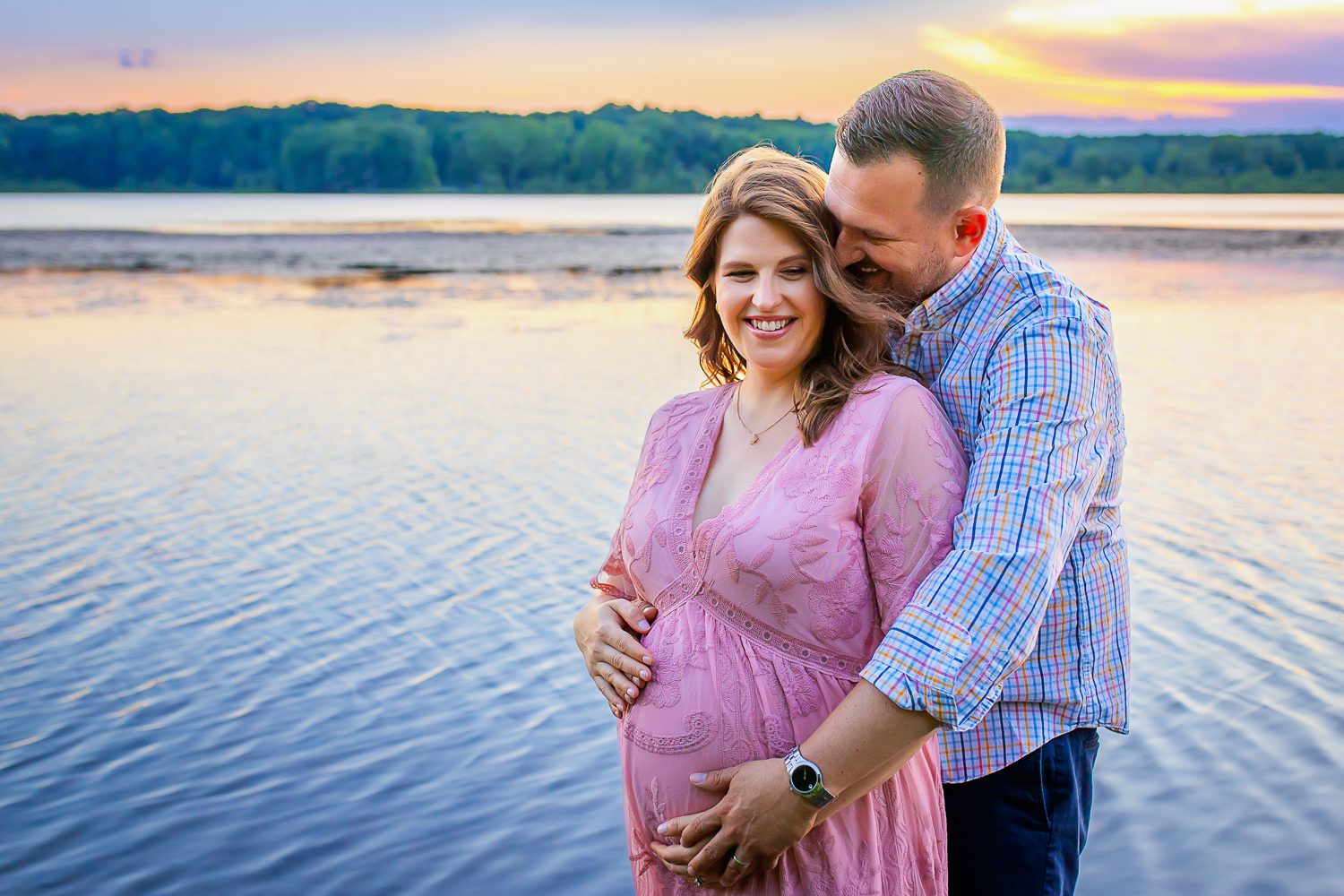 maternity photographer in rochester ny captures expectant parents smiling while the sun sets over the water